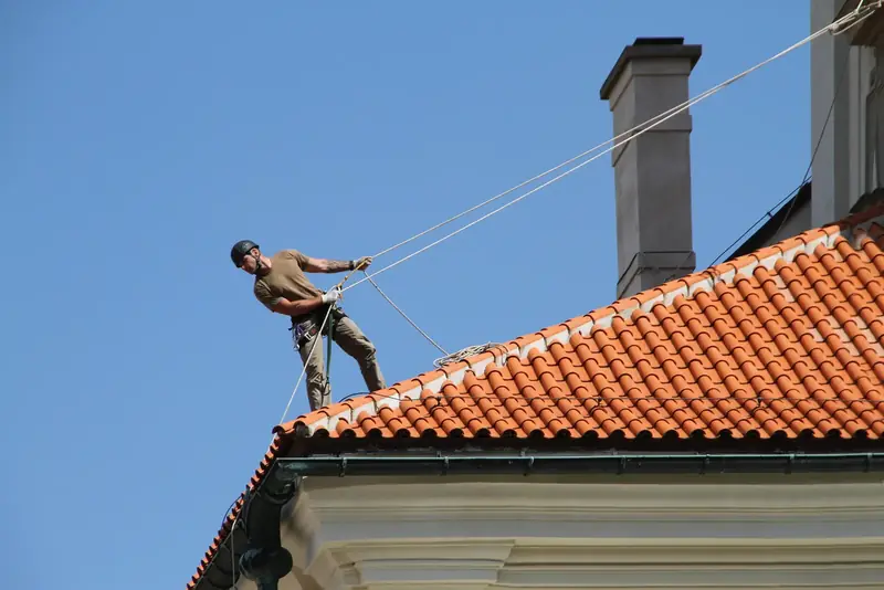 Couvreur encordé qui travaille debout sur un toit de tuiles rouges, sous un ciel bleu