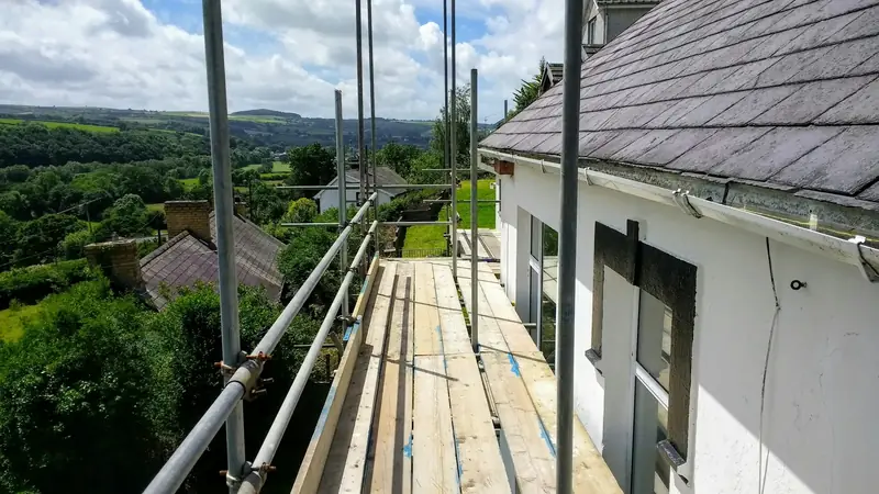 Échafaudage dressé le long d’une maison au toit d’ardoise, avec la campagne verdoyante en contrebas