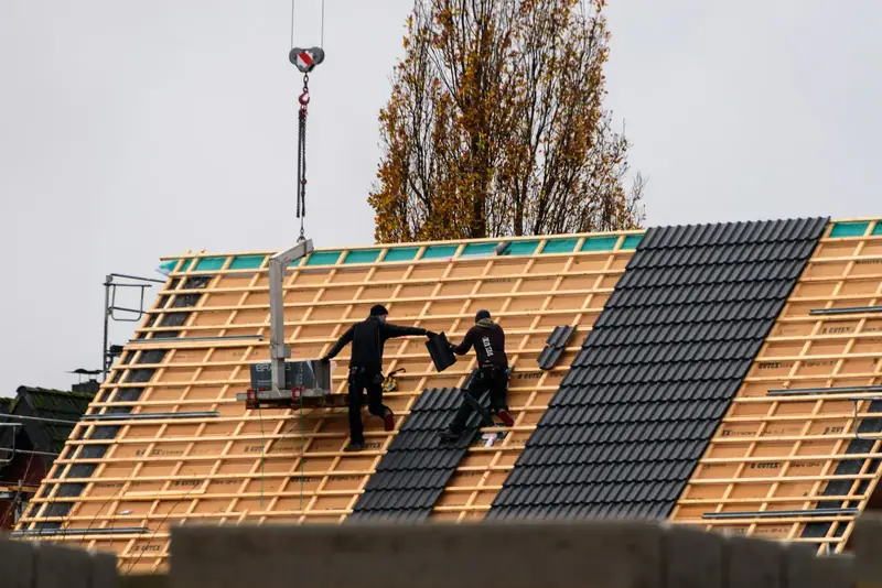 Deux couvreurs posent des tuiles sombres sur un versant neuf en liteaux, une grue au-dessus du chantier