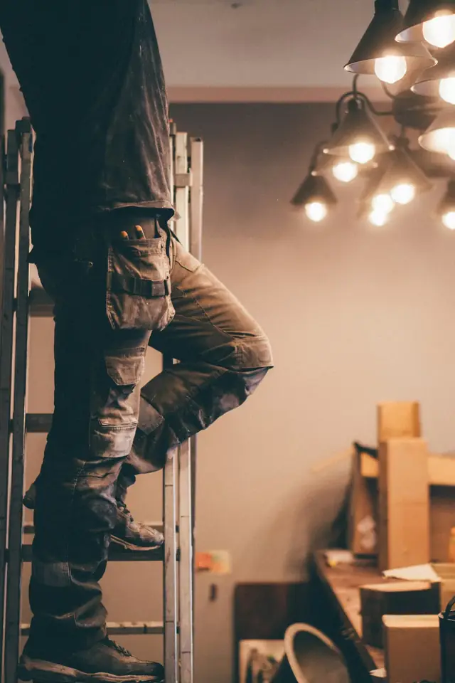Artisan sur un escabeau, vu au niveau des jambes, qui installe des luminaires dans une pièce en travaux