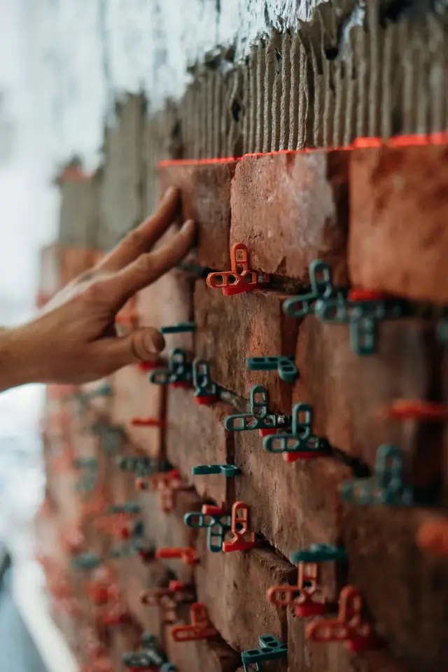 Mains d’un maçon qui alignent des briques rouges sur un mur à moitié monté, avec des cales